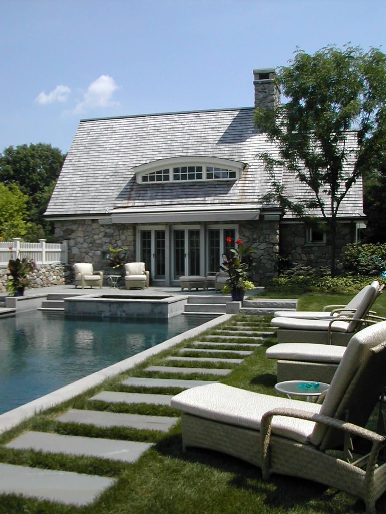 A hot tub and poolhouse anchor the far end of the swimming pool while the main house is on the opposite side. Widely-spaced slabs of bluestone surround the pool and are set into the lawn.