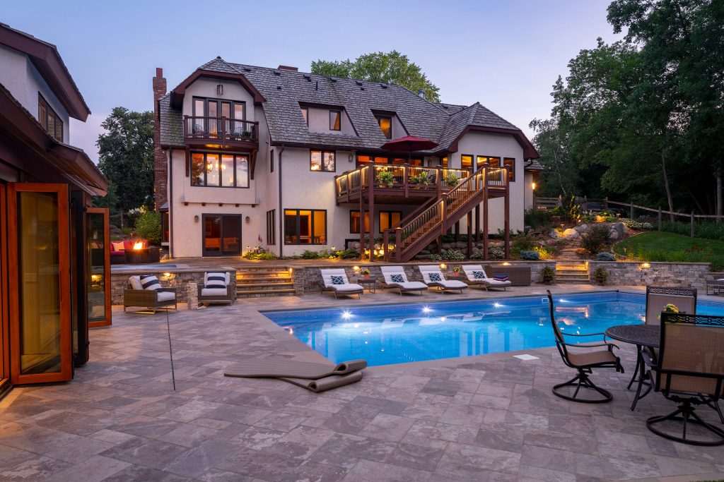 Backyard landscape with swimming pool, pool house, natural stone patio and warm lighting.