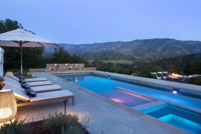 Colored pool lights accentuate pink moment of sunset over mountain views. Outdoor seating below pool with open firepit.