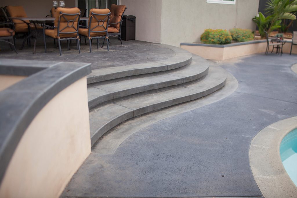 Dark stamped concrete steps down from the backyard patio area to the pool. Their curved design mimics the movement of the pool. TRU Landscape Services by TRU Landscape Services | Outdoor Kitchens | Backyard Design | Outdoor Living Spaces | Landscape Design