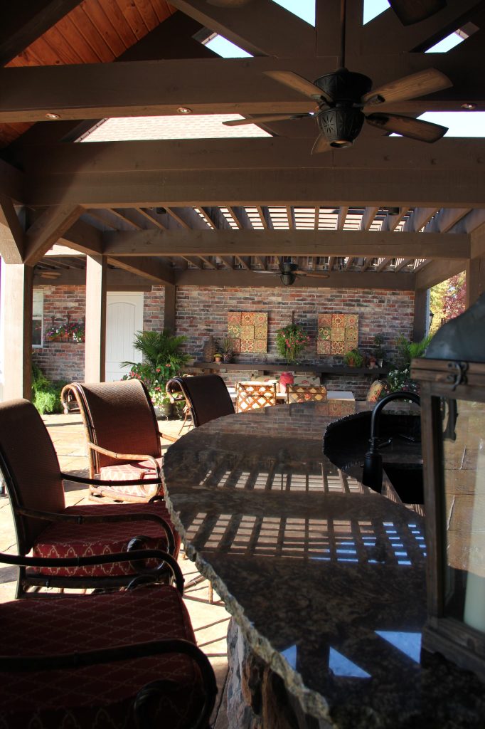 Outdoor Kitchen constructed with brick, stone, & timber framing with island seating and adjacent dining pergola that connects to rear porch. Photo by S Jasan