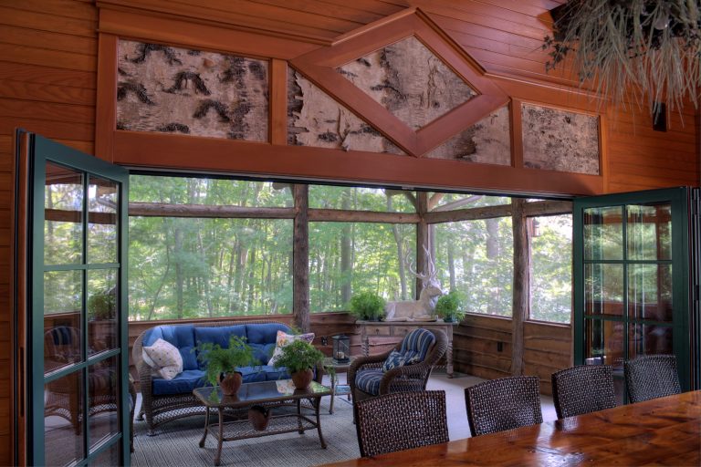Screened porch opens to living area via folding doors, that can close off the space in winter. Wicker, blue cushions and greenery complement the birch bark details