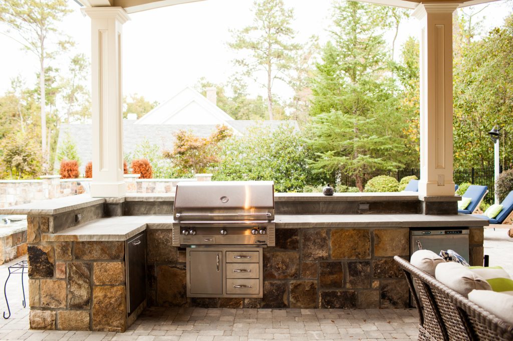 The L-shaped outdoor stone kitchen and bar area overlooks the pool and features a wraparound raised bar with seating for more than 10 guests and contains a grill, refrigerator, storage doors, and ice maker. by Boyce Design and Contracting | Outdoor Kitchens | Backyard Design | Outdoor Living Spaces | Landscape Design