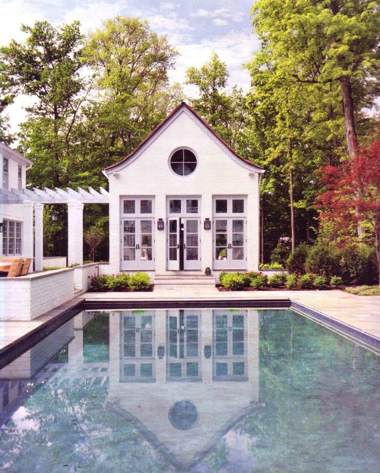 The pool house is connected to the main house by an open pergola. The gently sloping roof gives a whimsical appearance to the building and the white brick reflects in the soft turquoise blue of the pool.