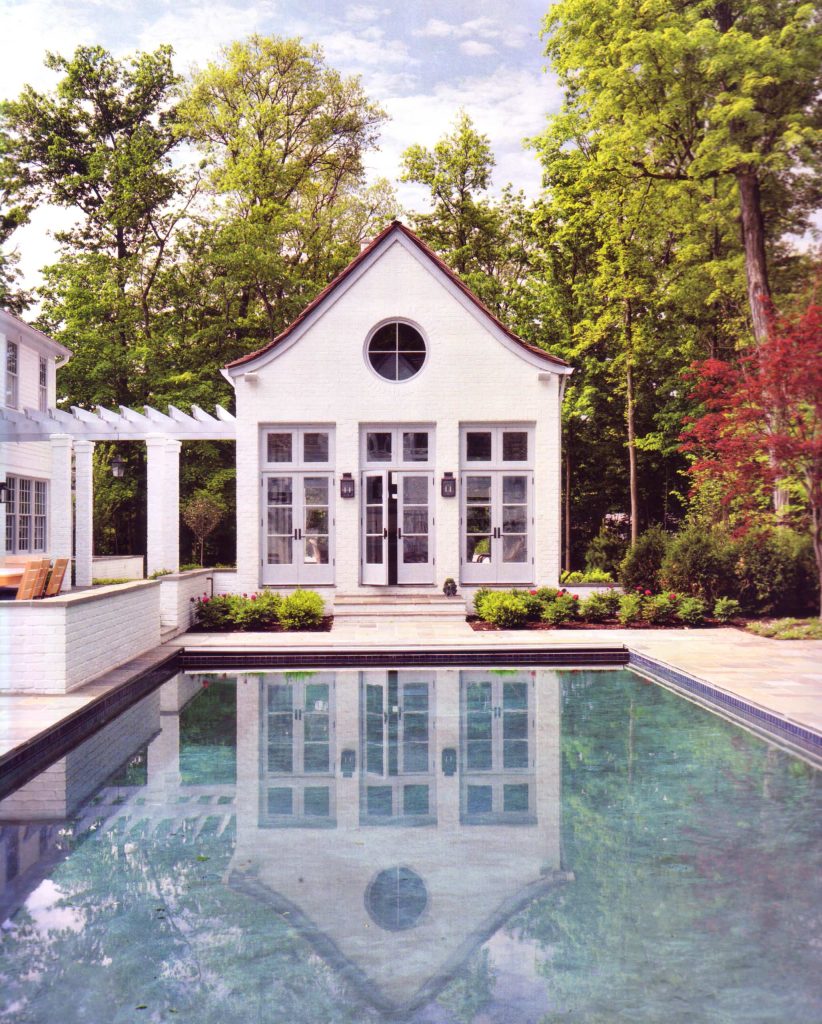 The pool house is connected to the main house by an open pergola. The gently sloping roof gives a whimsical appearance to the building and the white brick reflects in the soft turquoise blue of the pool.