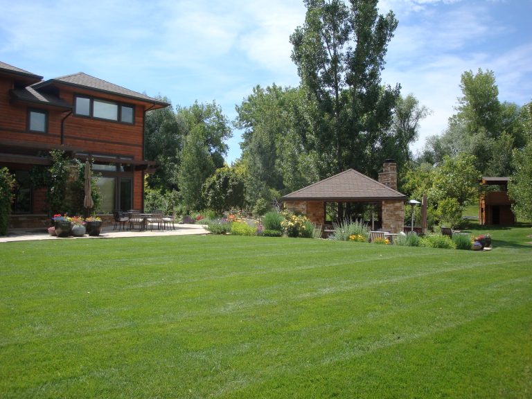 This backyard is open and inviting to run around or relax by the kitchen area, or any of the other decorated seating. Flowerbeds outline the kitchen area, and trees invoke the yard in meticulous spots.