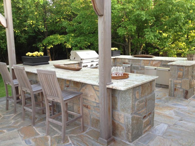 This large open outdoor kitchen provides ample space to hold food and beverages during get-togethers. The granite countertop matches well with the color of the pergola and stone.