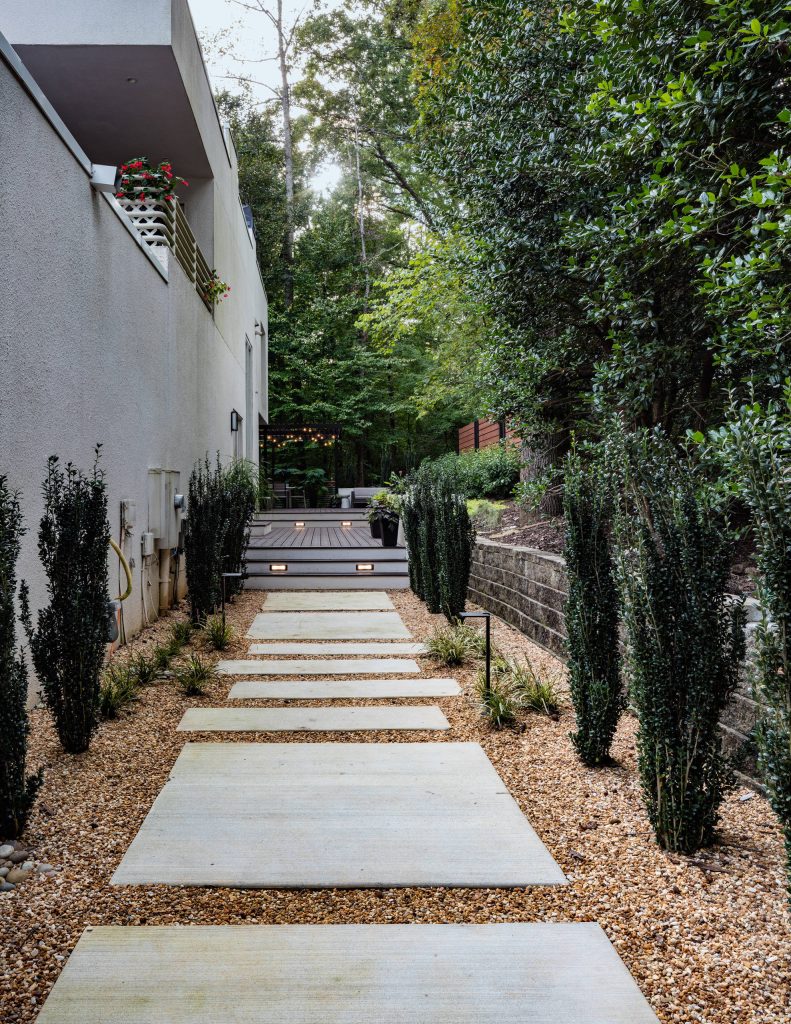 Custom decorative concrete pavers surrounded by pebble stone line the entrance to this outdoor space. The modern landscaping composed of tight, linear plantings with hues of green provides privacy from neighboring properties. The deck steps span the entire width of the deck and have built-in recessed lighting so that the space is well-defined in the evening as well as the daytime.