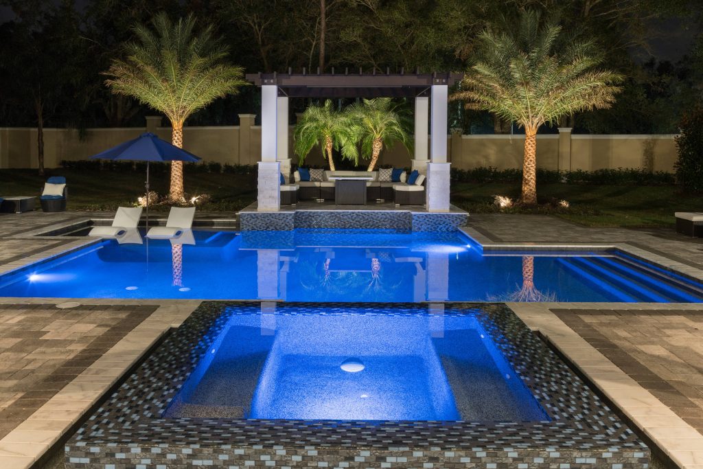 This resort style pool has a pergola canopy over the seating area and fire table. The pool has glass mosaic tiles and a spa with a shallow sundeck and plenty of space for lounge chairs and umbrellas. This Florida Modern Home in Lake Mary is designed and built by Orlando Custom Home Builder Jorge Ulibarri, a top custom builder in Orange, Seminole, Lake County, Orlando, Winter Park, Maitland, Lake Mary, Lake Nona, Sanford, Mt. Dora, Longwood, Altamonte Springs, and Windermere. Jorge Ulibarri custom homes is proficient in designing all styles of homes including modern, classical, traditional, Tuscan, and Spanish Mediterranean. For more go to https://cornerstonecustomconstruction.com