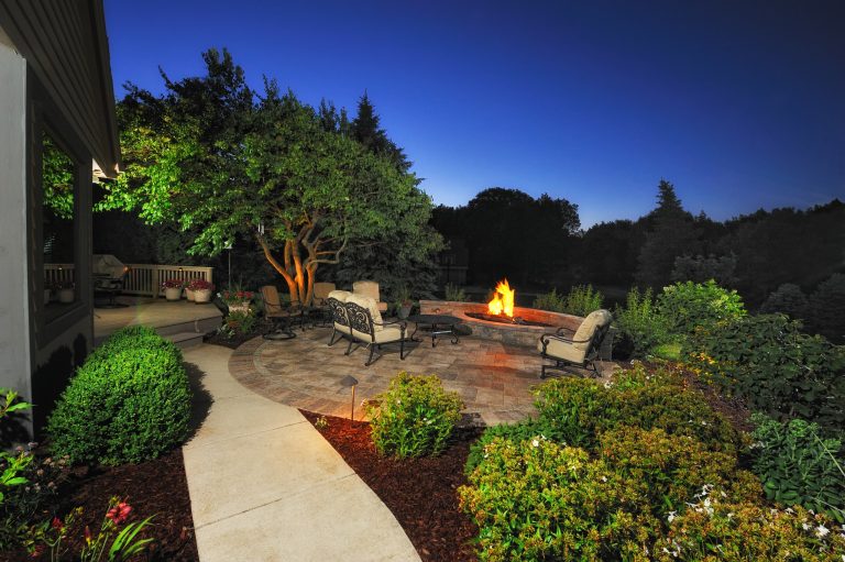 The gas fire pit casts a warm glow to the landscape as if it was always there. LED lighting as path lights and wall lights illuminate the paving, while a Pekin Lilac tree is illuminated by an uplight.