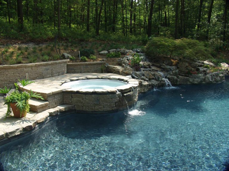 This 22-year-old gunite pool needed a facelift. A nonfunctioning waterfall, uncomfortable spa and grass as its patio were the reasons the client called Bill Renter from the Deck & Patio Company of Huntington Station New York. The first step was to demolish the existing spa, waterfall, wood retaining wall and rock coping. Next, the new acrylic spa was installed. A seven foot high moss rock waterfall and boulder coping were installed beautify and retain the sloping woodland behind the pool. Because of the natural setting of the pool, careful consideration was given to the patio material. The designers suggested a tumbled pavingstone and wall system called techno-bloc. The colors of the patio were a blend of a tan color and a natural grey color to tie in with colors of the boulders used. The patio has the look of natural stone, but it is cool even on hot summer days and has the durability of a paver patio. This new technology in the paving and matching wall systems insures a lifetime of easy car for these surfaces. The final touch was the colorful landscape which is beautiful despite the challenge of a mostly shady woodland setting. http://www.deckandpatio.com/DP_Blog/?p=1182 Deck and Patio Company www.deckandpatio.com