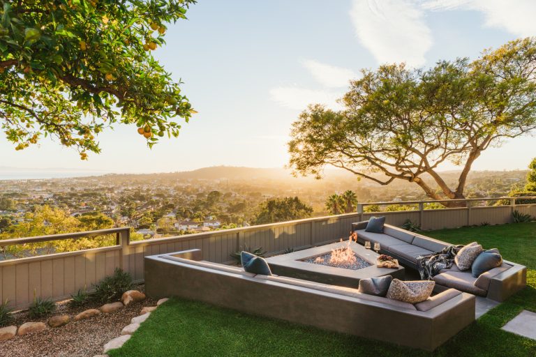 Modern built-in concrete seating area includes custom made couch cushions and a geometric fire pit. Surrounded by synthetic grass and flagstone, this side yard uses neutral colors to highlight the Santa Barbara view. Waterwise landscaping surrounds the yard. Stone edging is used to separate turf and gravel.