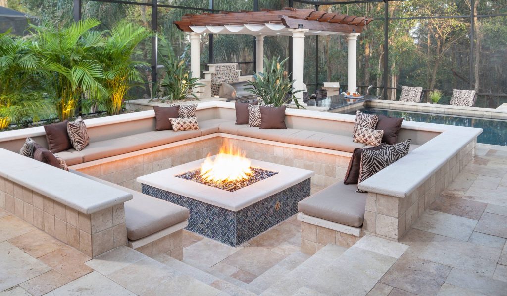 Adjacent to the television lounge is a recessed seating nook with a large stone fire pit. The uniquely designed seating delivers intimacy and comfort. with a cushioned surrounding bench adorned with custom throw pillows. The fire pit is the heart of this area providing warmth and gathering charm. by Ryan Hughes Design/Build | Fire Pits|Backyard Design|Outdoor Living Spaces|Landscape Design|Backyard Ideas|Landscaping|Landscaping Ideas|Landscape Installation|Fire Feautures|Fire Pit
